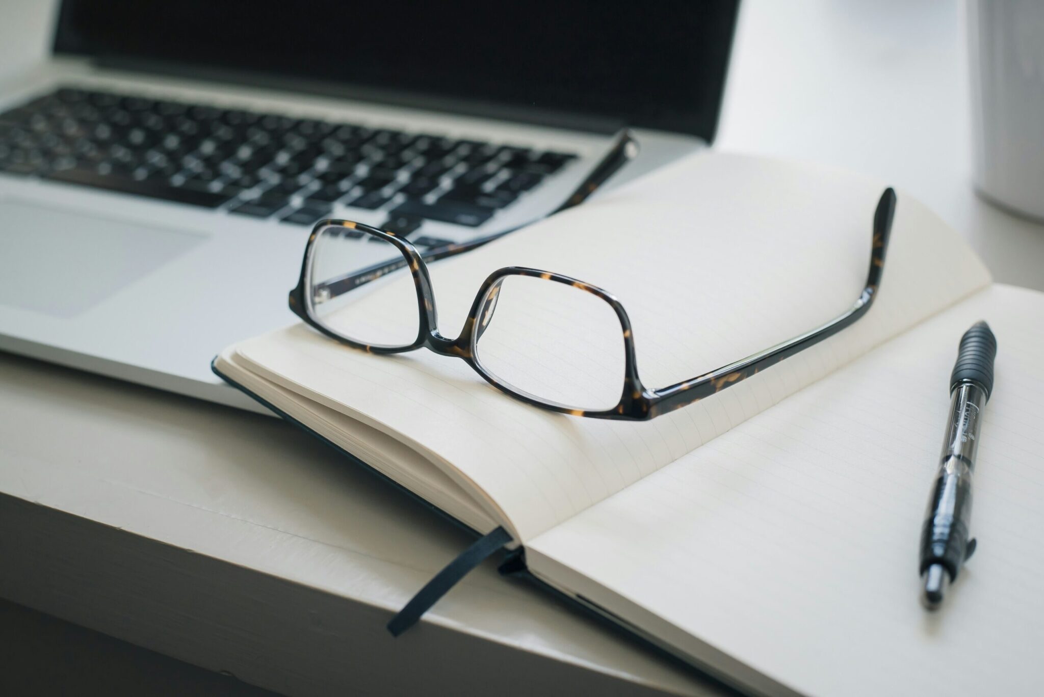 Close-up of a workspace featuring a pair of tortoiseshell eyeglasses resting on an open lined notebook. A black pen lies beside the notebook, and a laptop is partially visible in the background.