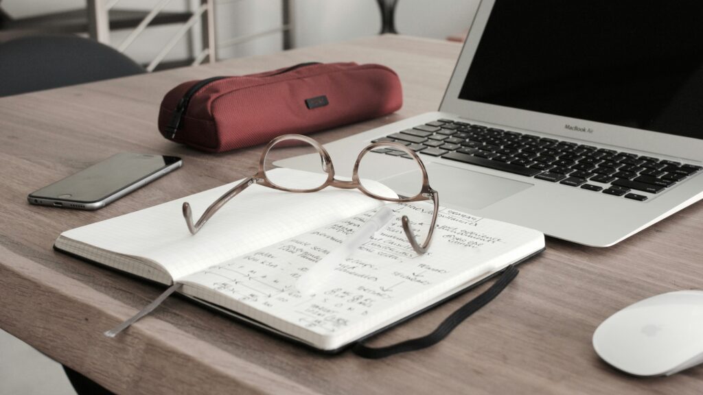 Workspace setup on a wooden desk featuring an open notebook with handwritten notes, a pair of round eyeglasses resting on top, a white pen, a closed red pencil case, a MacBook Air laptop, a smartphone, and an Apple Magic Mouse.