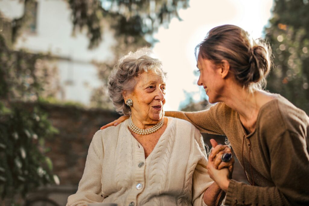 Joyful adult daughter embracing and surprising her smiling senior mother in a garden, conveying warmth, connection, and multi-generational family bonding.